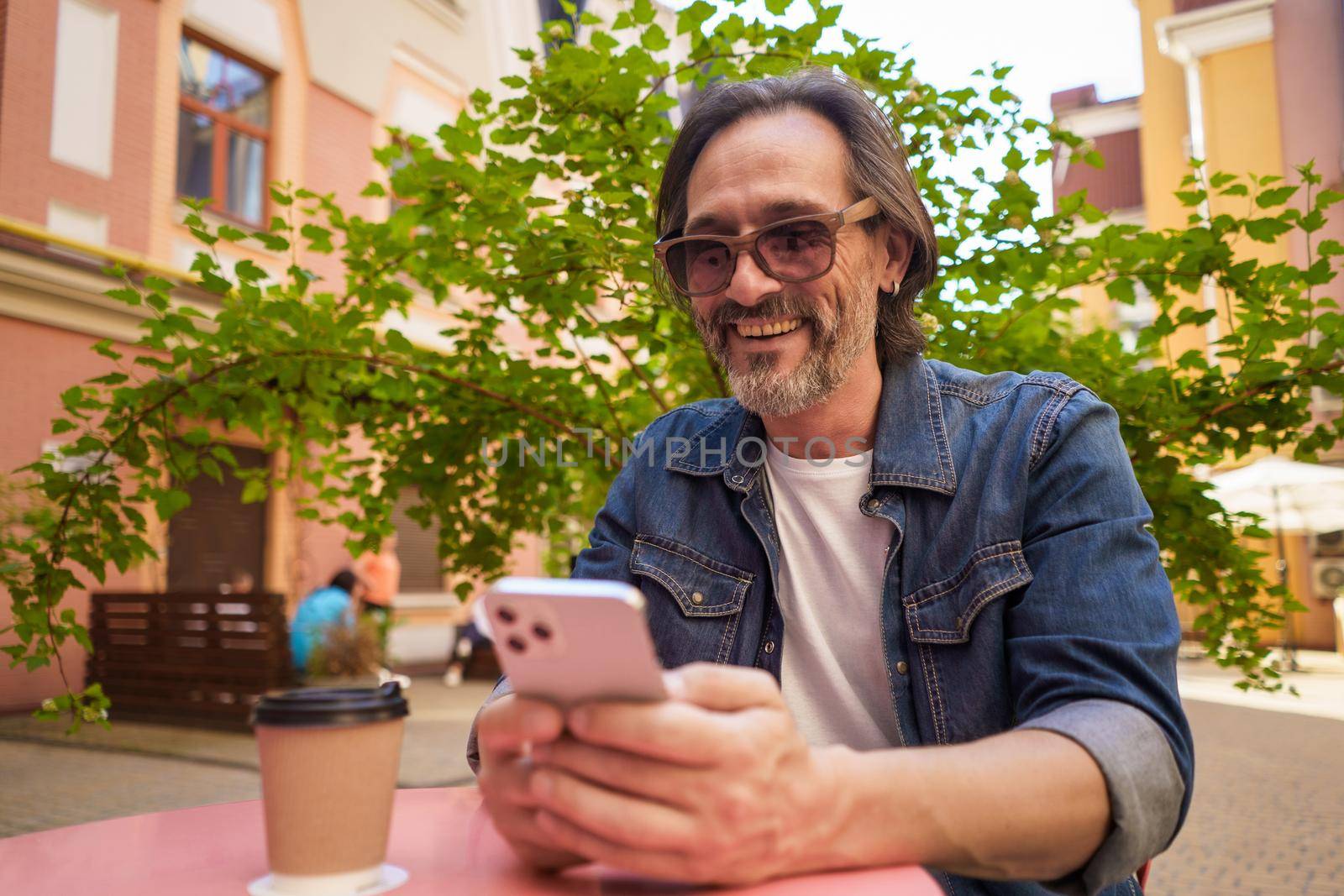 Middle aged handsome freelancer man texting or having a work video call while enjoying coffee outdoors. Mature man looking at phone in hands sitting at cafe while working outdoors.