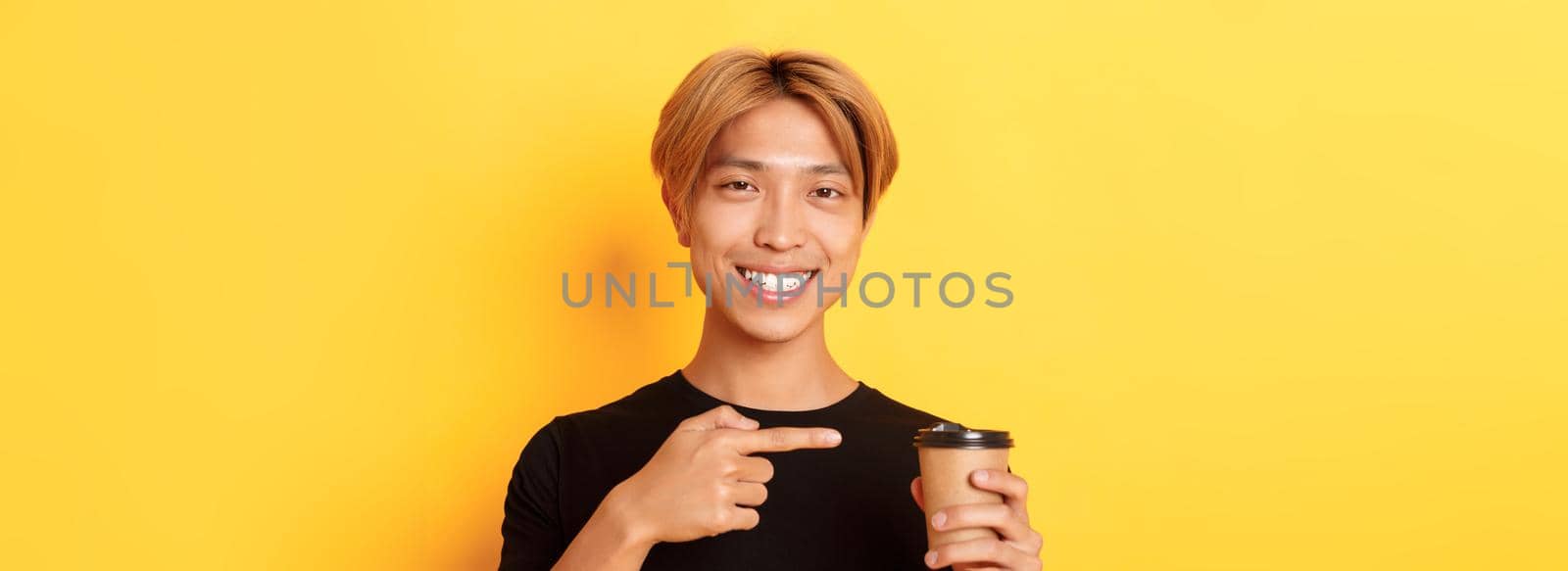 Close-up of satisfied handsome asian blond guy, smiling delighted and pointing finger at delicious cup of coffee, standing over yellow background by Benzoix