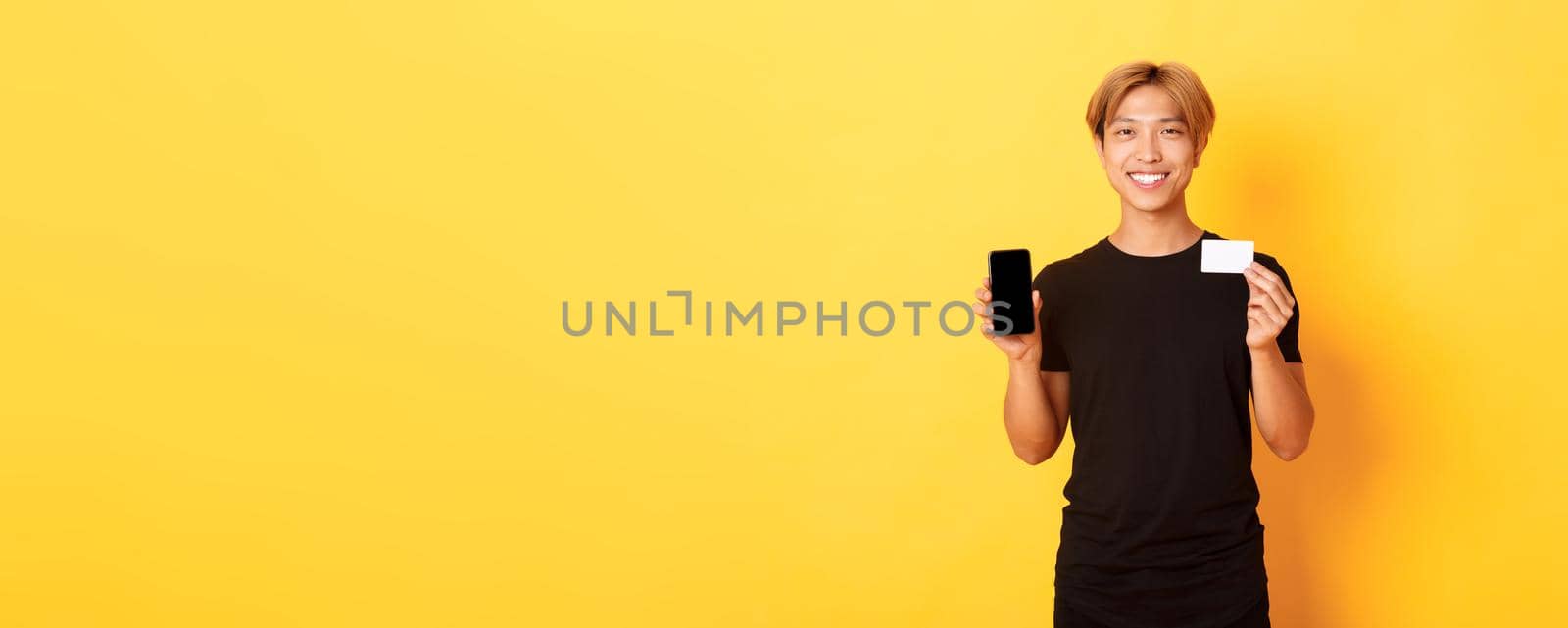 Portrait of handsome young asian guy showing smartphone screen, banking app and credit card, standing yellow background and smiling by Benzoix