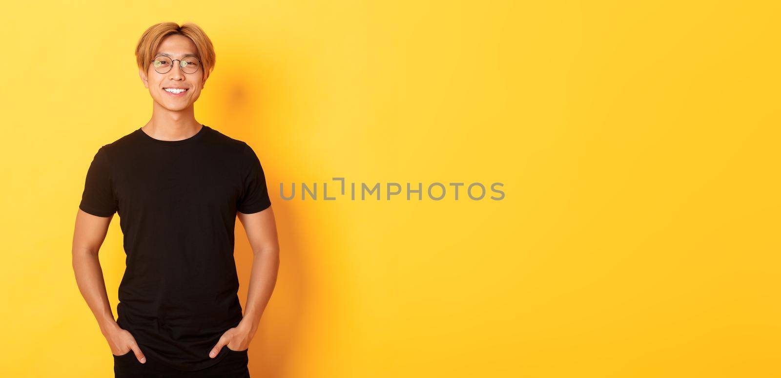 Handsome asian blond guy in glasses standing in black clothes and smiling pleased, yellow background by Benzoix