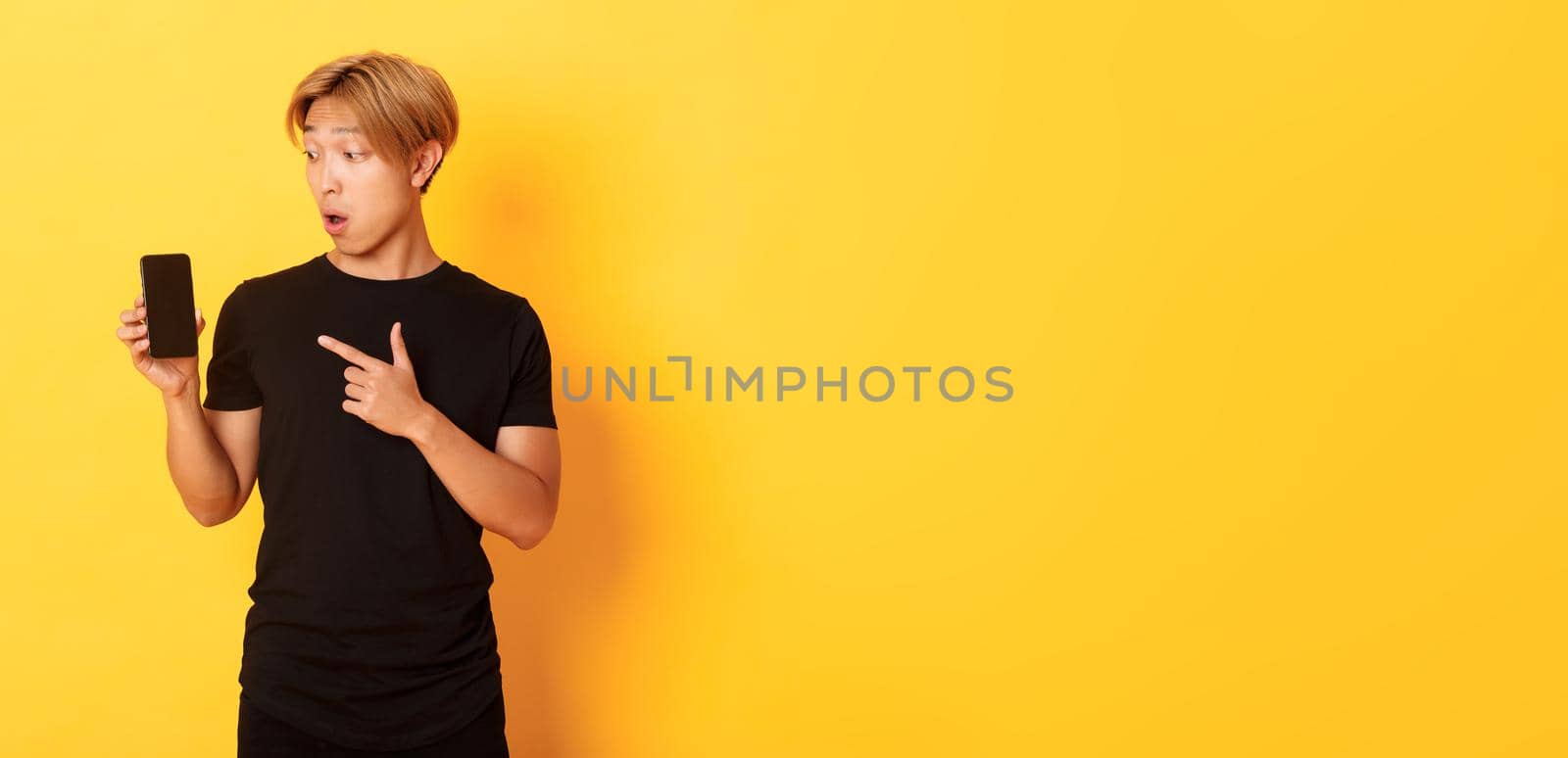 Portrait of handsome asian man showing something on smartphone screen, pointing at mobile phone display, standing over yellow background by Benzoix