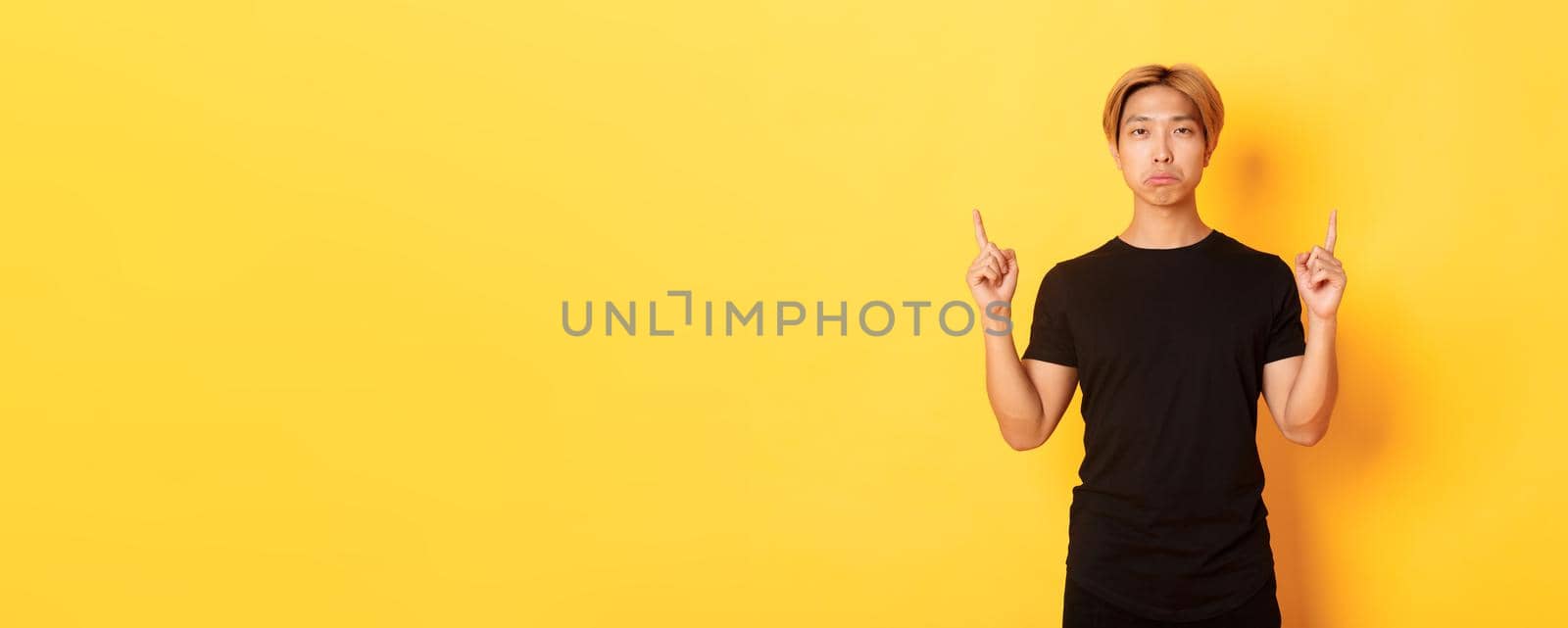 Portrait of sulking gloomy asian guy looking disappointed, pointing fingers up, yellow background.