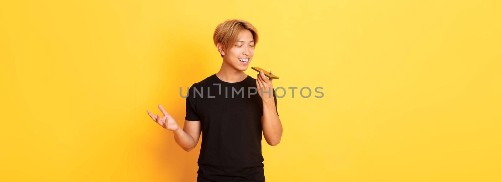 Portrait of stylish handsome korean guy with blond hair record voice message on mobile phone, holding smartphone near mouth and speaking, standing yellow background.