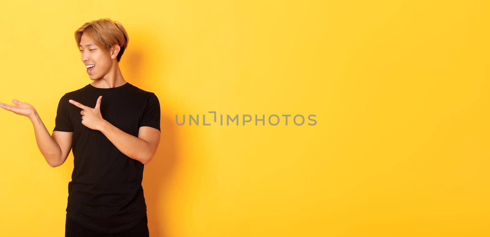 Portrait of handsome smiling asian guy holding something on hand and pointing finger at it, standing yellow background pleased.