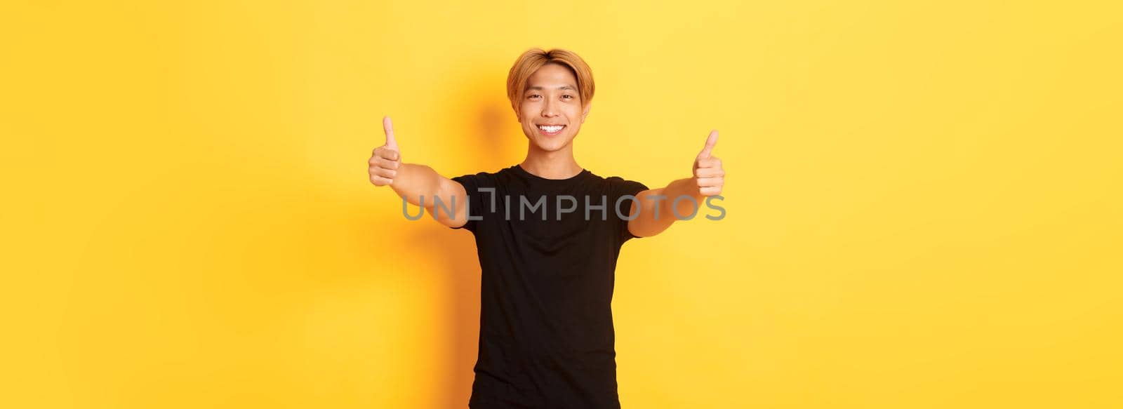 Portrait of assured handsome asian man showing thumbs-up in approval, like idea, standing yellow background.