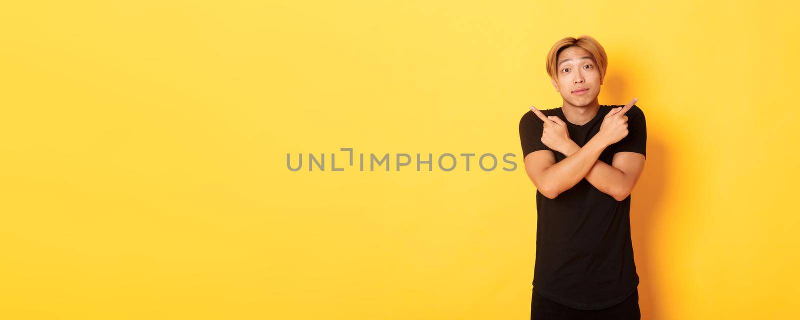 Indecisive handsome guy shrugging puzzled and pointing fingers sideways, standing yellow background.