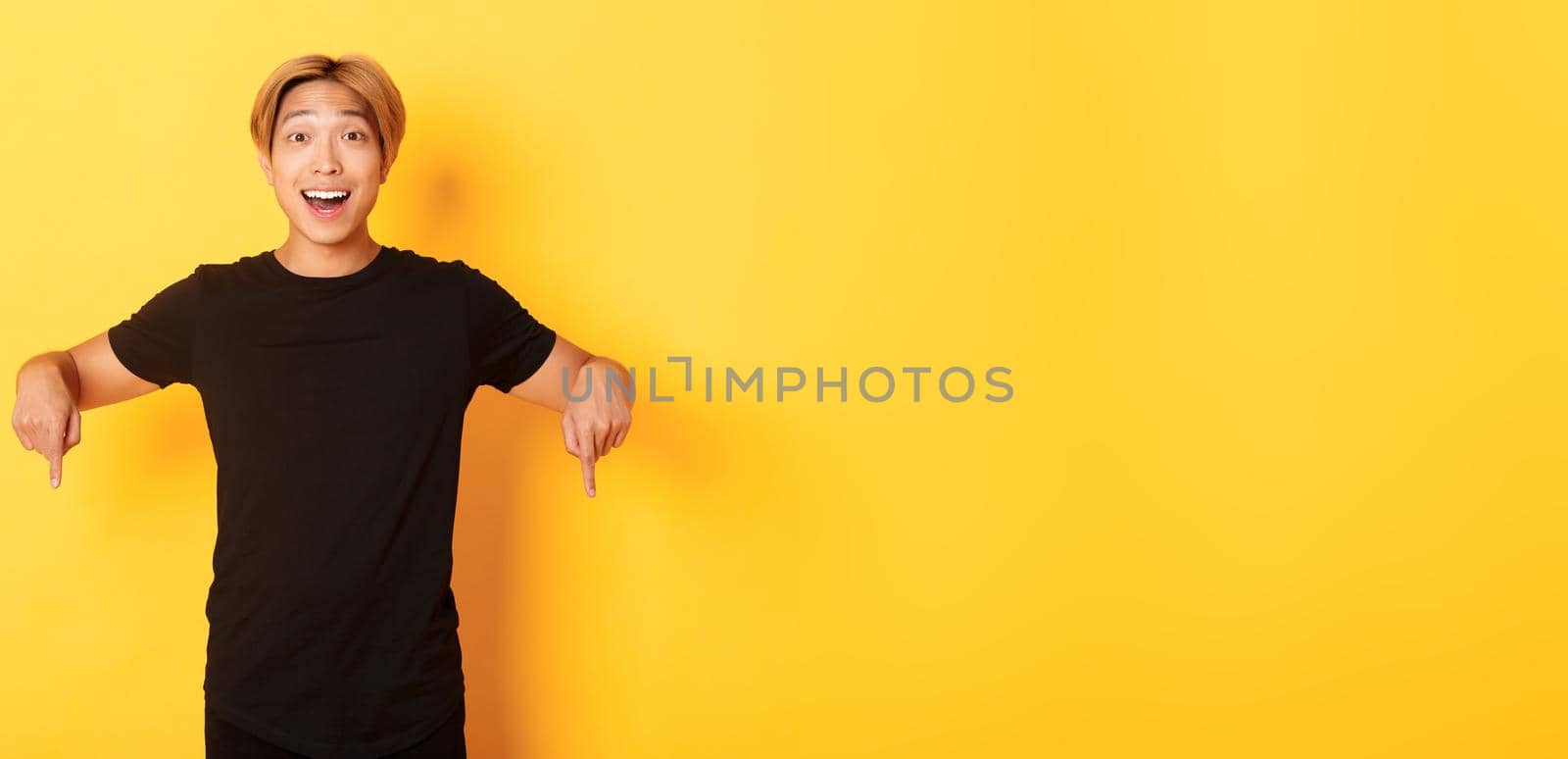 Portrait of excited smiling asian guy with blond hair, pointing fingers down, showing banner and looking happy, yellow background.
