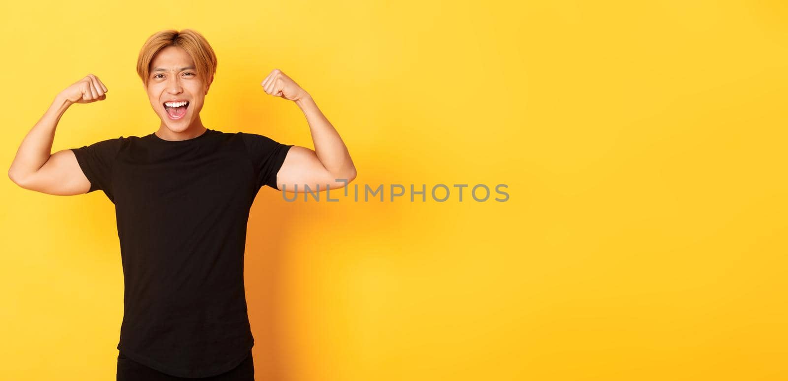 Portrait of happy and confident asian blond guy, flexing biceps, showing strong big muscles, standing yellow background by Benzoix