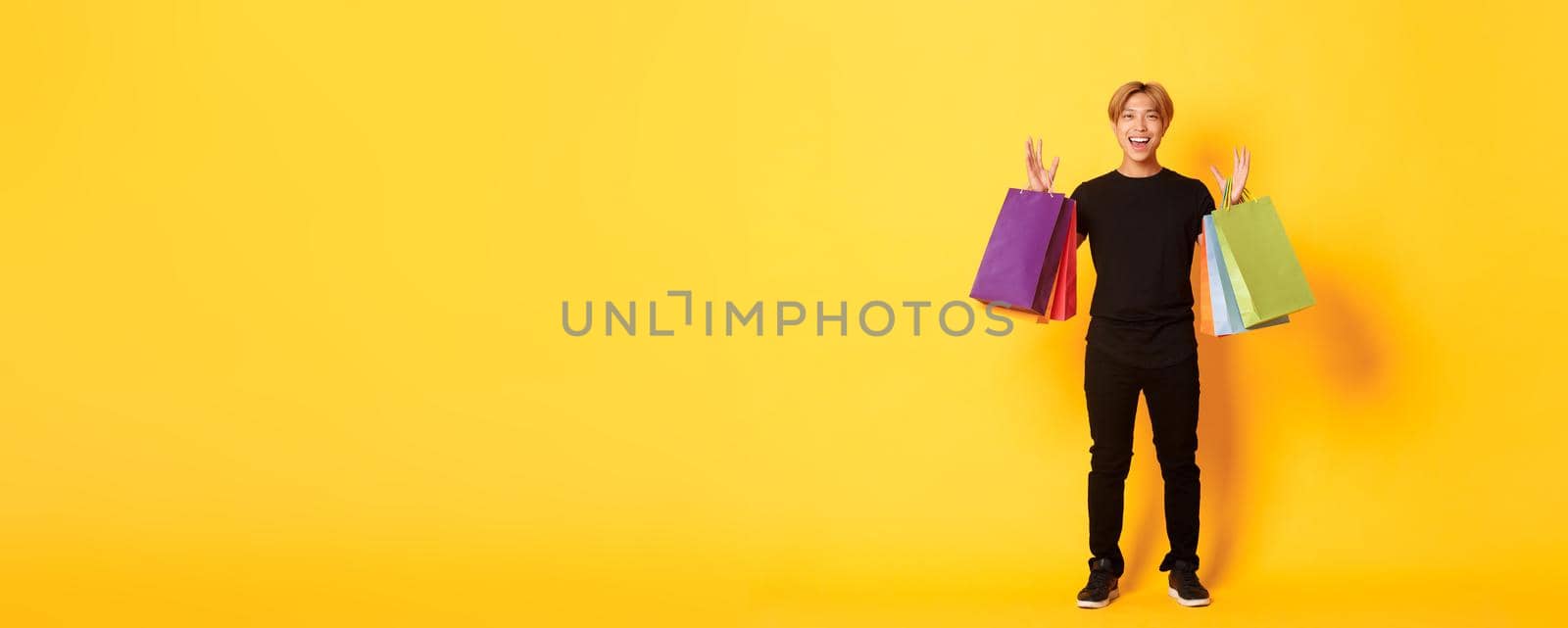 Full-length of happy handsome asian guy on shopping, holding bags and smiling, yellow background.