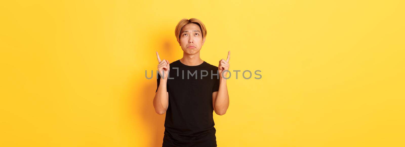 Portrait of gloomy asian blond man, wearing black t-shirt, sulking disappointed and pointing fingers up, yellow background.