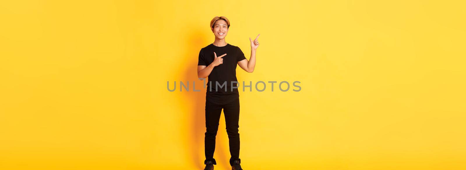 Full-length of smiling attractive korean guy pointing fingers left, showing logo, yellow background.