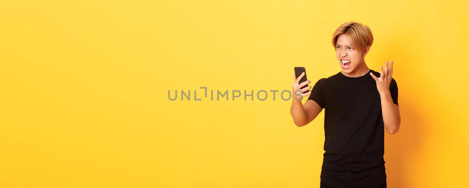 Portrait of angry and pissed-off asian man looking mad at smartphone screen, having argument during video call, standing yellow background.