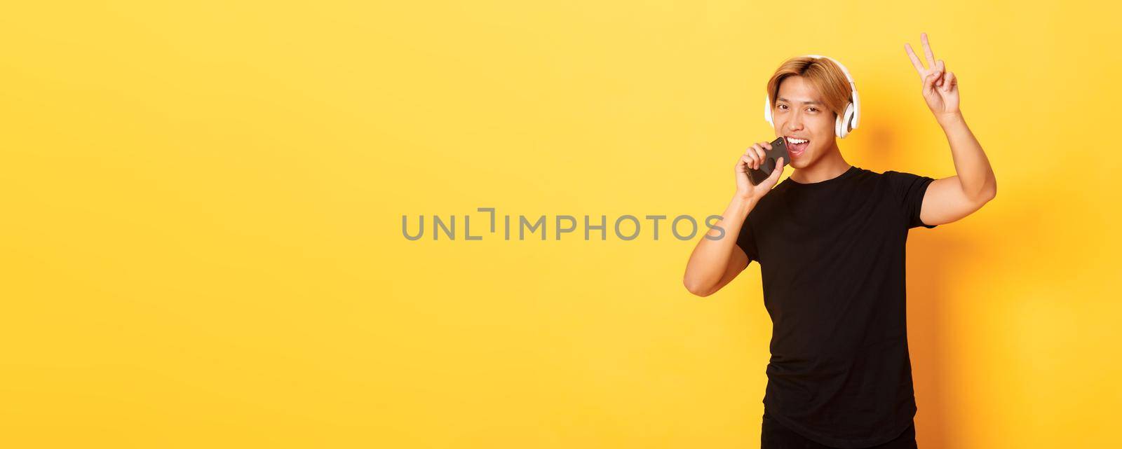 Sassy handsome asian guy in headphones, playing karaoke app, singing into mobile phone microphone, showing peace gesture, standing yellow background.