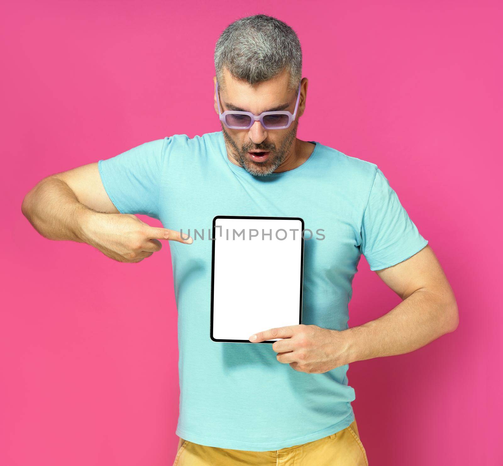 Pointing at digital tablet handsome man looking at white screen lowered his head wearing casual blue shirt and sunglasses isolated on pink background. Mobile app advertisement by LipikStockMedia