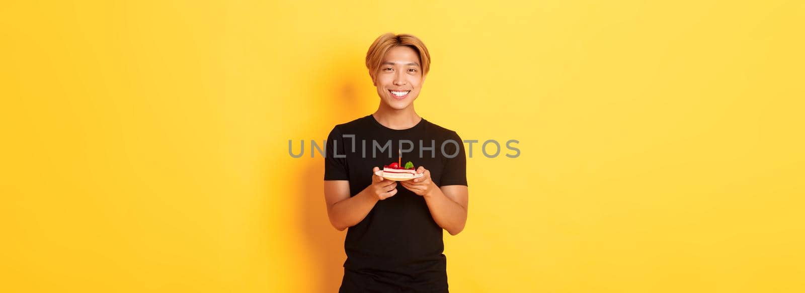 Handsome happy asian blond guy, smiling pleased as celebrating birthday, holding b-day cake, standing over yellow background by Benzoix