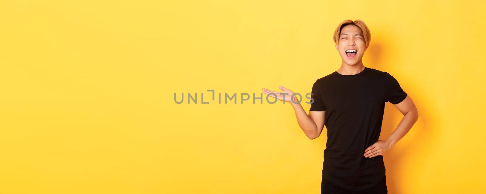 Portrait of happy blond asian guy, winking sassy and smiling, holding something on hand over yellow background.
