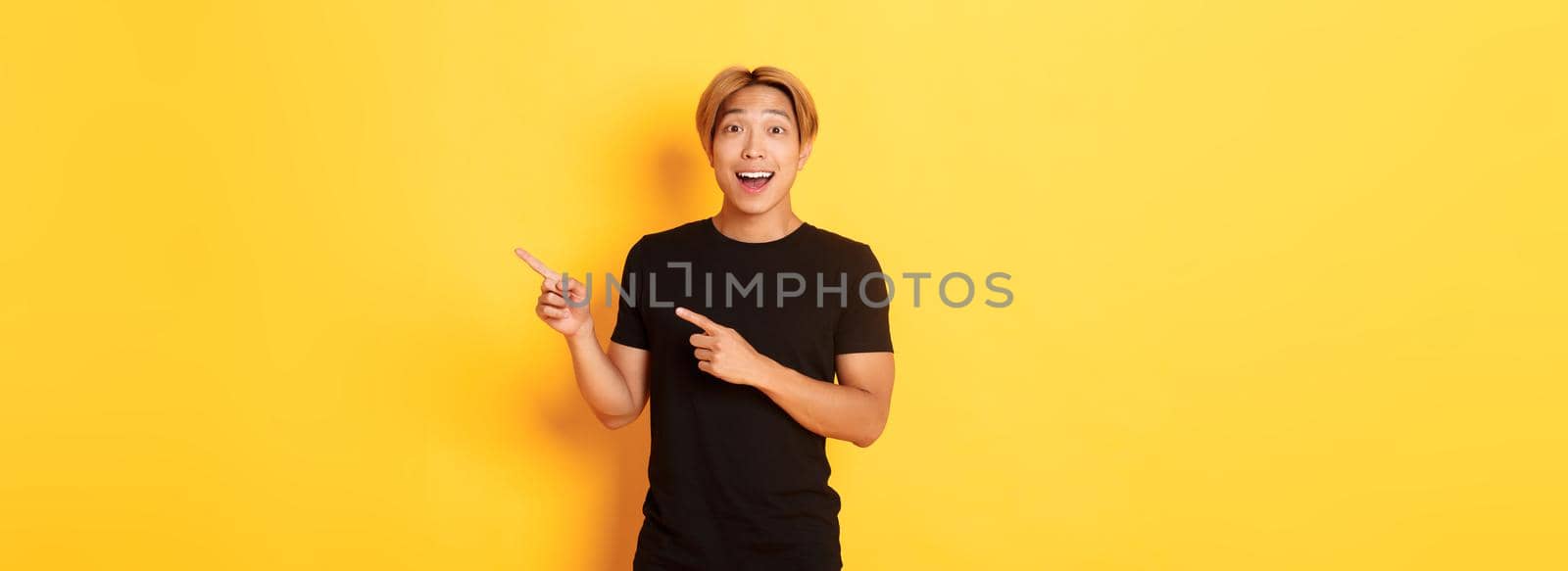 Amazed handsome korean guy, smiling fascinated and pointing fingers right, showing way, standing yellow background by Benzoix