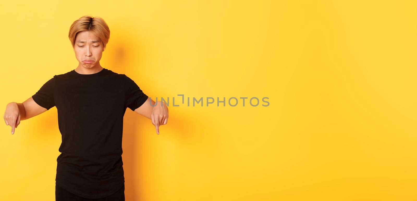 Portrait of gloomy and upset asian man sulking with regret, pointing and looking down at banner, yellow background by Benzoix