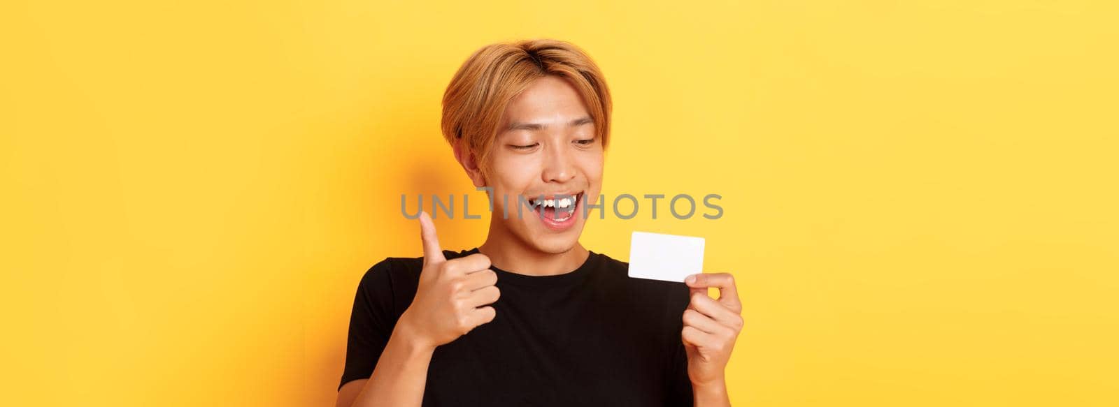 Close-up of happy and satisfied asian handsome guy, showing credit card and thumbs-up in approval, smiling amazed, standing yellow background by Benzoix