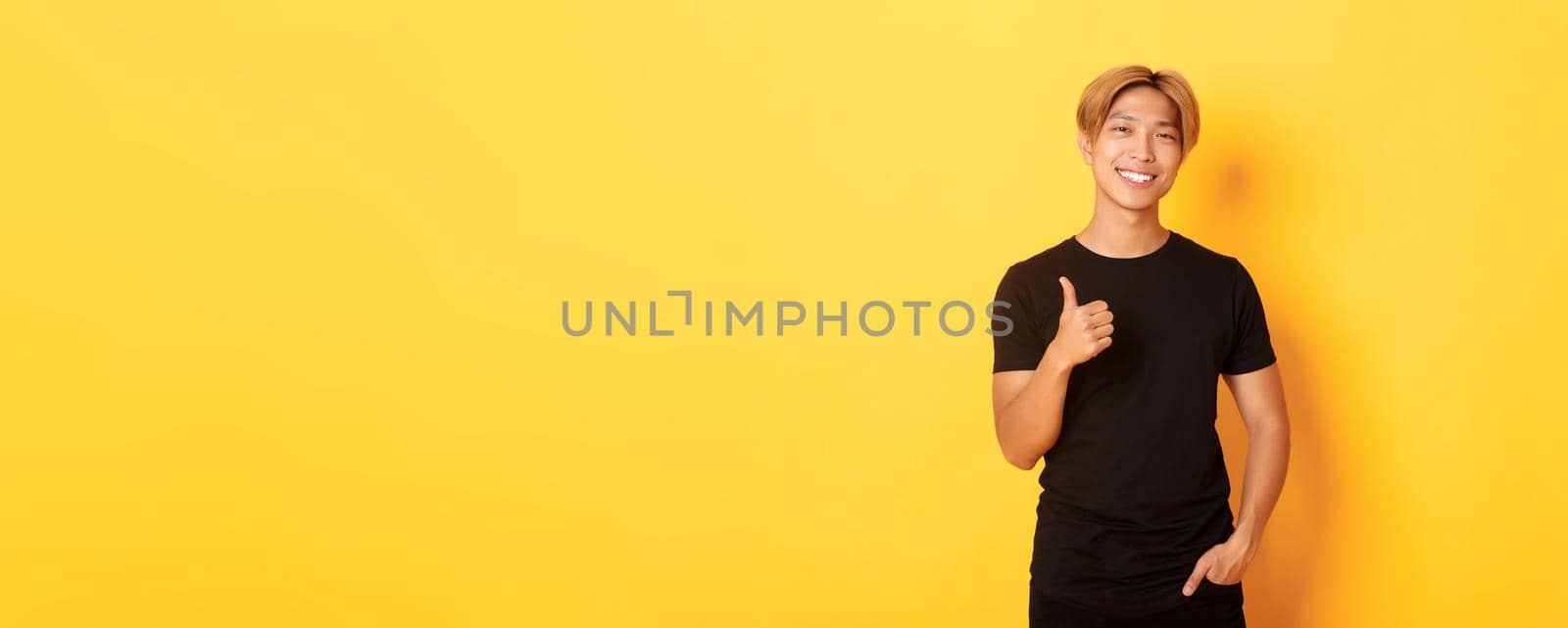 Portrait of satisfied handsome asian blond guy, smiling pleased, showing thumbs-up in approval, standing yellow background by Benzoix