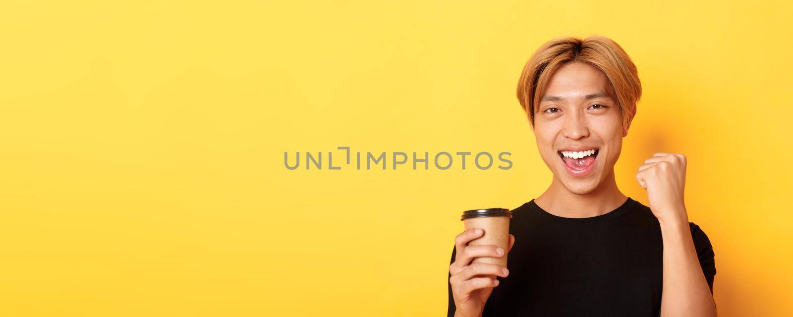 Close-up of energized handsome asian guy fist pump joyfully while drinking coffee, smiling excited over yellow background.