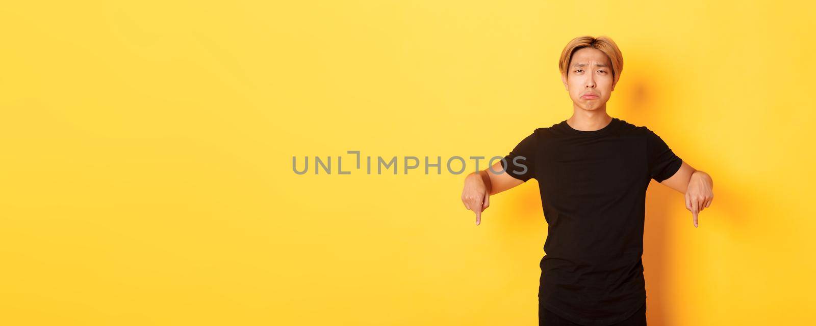 Portrait of disappointed gloomy asian guy, pouting upset and pointing fingers down with regret and uneasy feeling, yellow background.