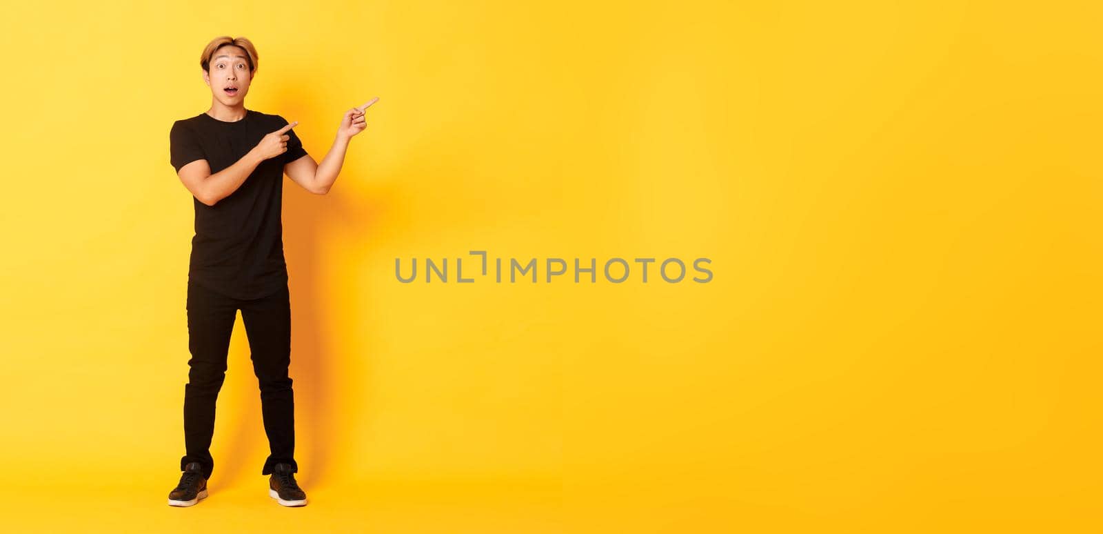Full-length of amazed and impressed attractive asian guy pointing fingers upper right corner, showing logo, yellow background.