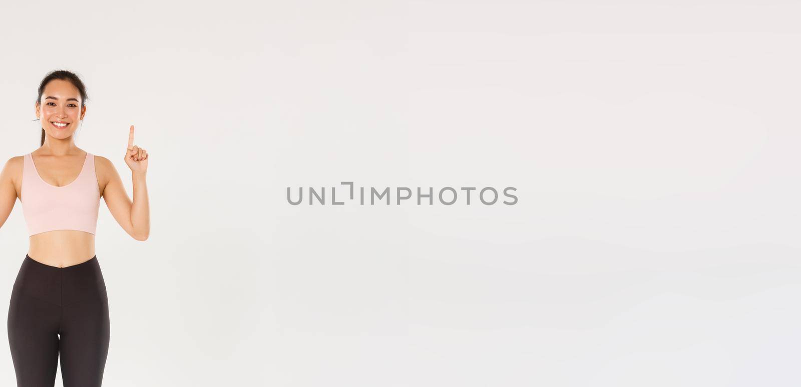 Sport, gym and healthy body concept. Full length of cheerful smiling and cute fitness girl, pointing fingers up, showing workout equipment advertisement, buy membership for training.