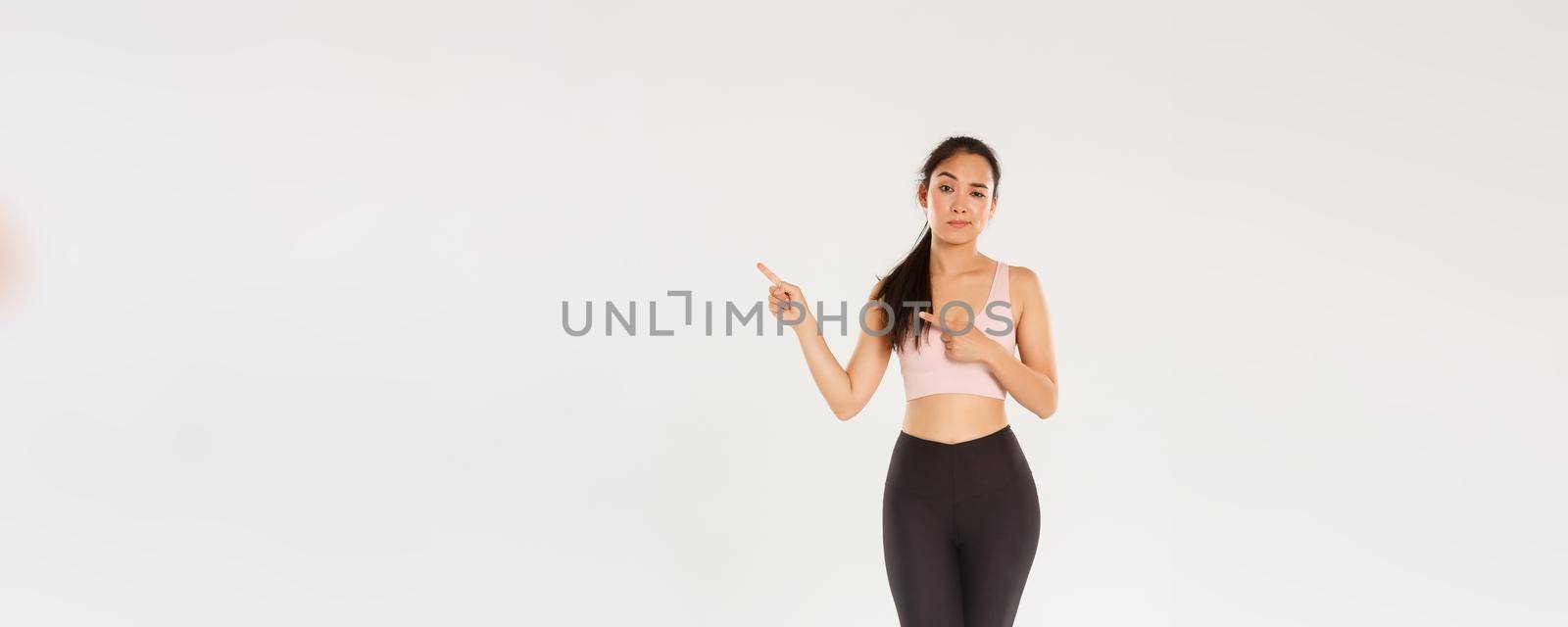 Full length of skeptical and doubtful asian female brunette, girl athlete have doubts, pointing fingers upper right corner, frowning and looking with disbelief, sportswoman showing promo.