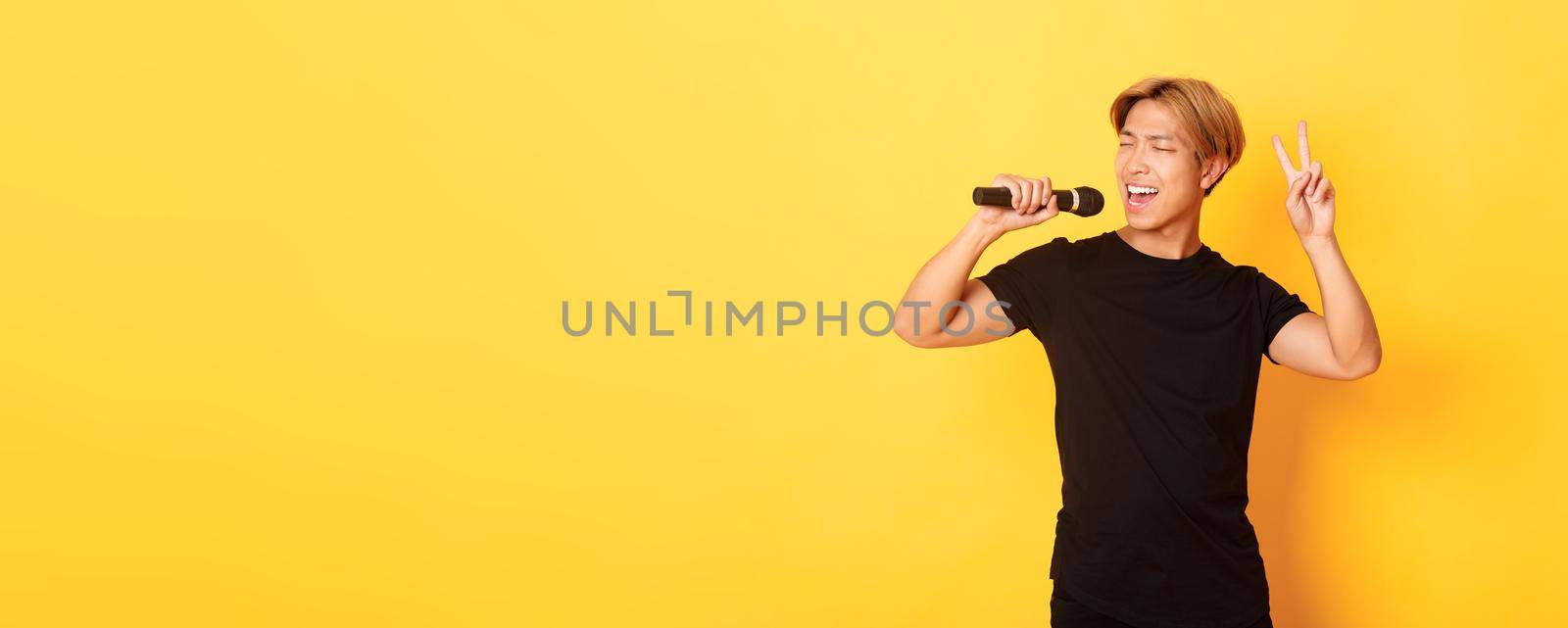 Carefree handsome asian guy performing song, singing into microphone and showing peace gesture, standing yellow background.
