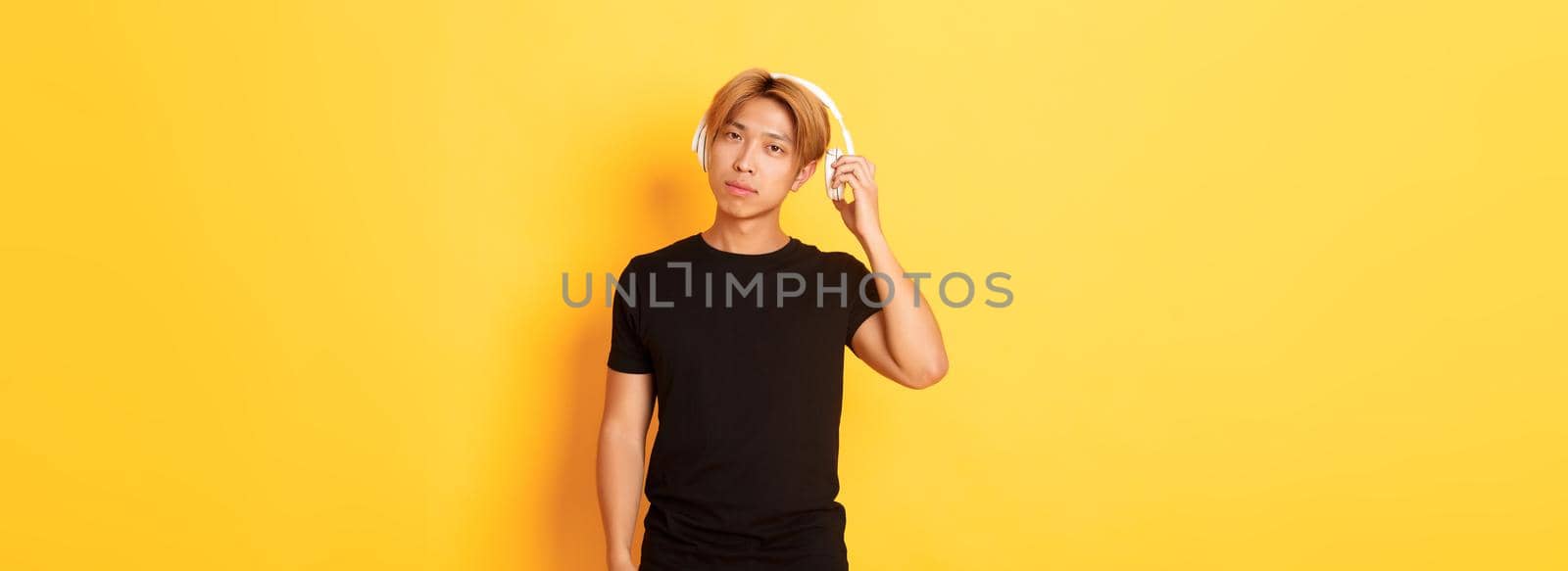 Young handsome asian guy with blond hair, take-off headphones to listen you, standing yellow background.