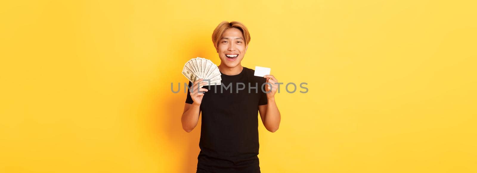 Young happy asian man with blond hair, showing credit card and money, smiling cheerful, standing yellow background.