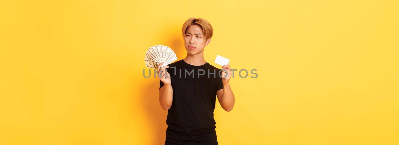 Indecisive and confused young asian guy looking puzzled at money while holding cash and credit card, yellow background.