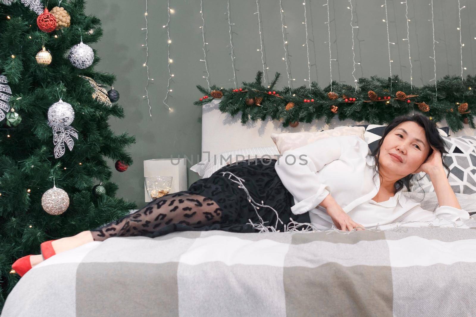 Senior asian full length woman rest on bed with xmas decoration and lights, portrait of attractive elderly smiling lady dreaming and relaxing at home at christmas holiday