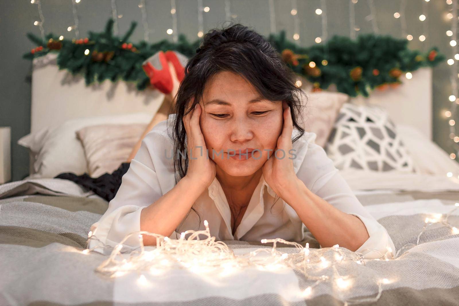 Senior asian woman relax on bed looking at xmas lights, portrait of attractive elderly smiling lady in bedroom at home at christmas holiday, atmospheric mood