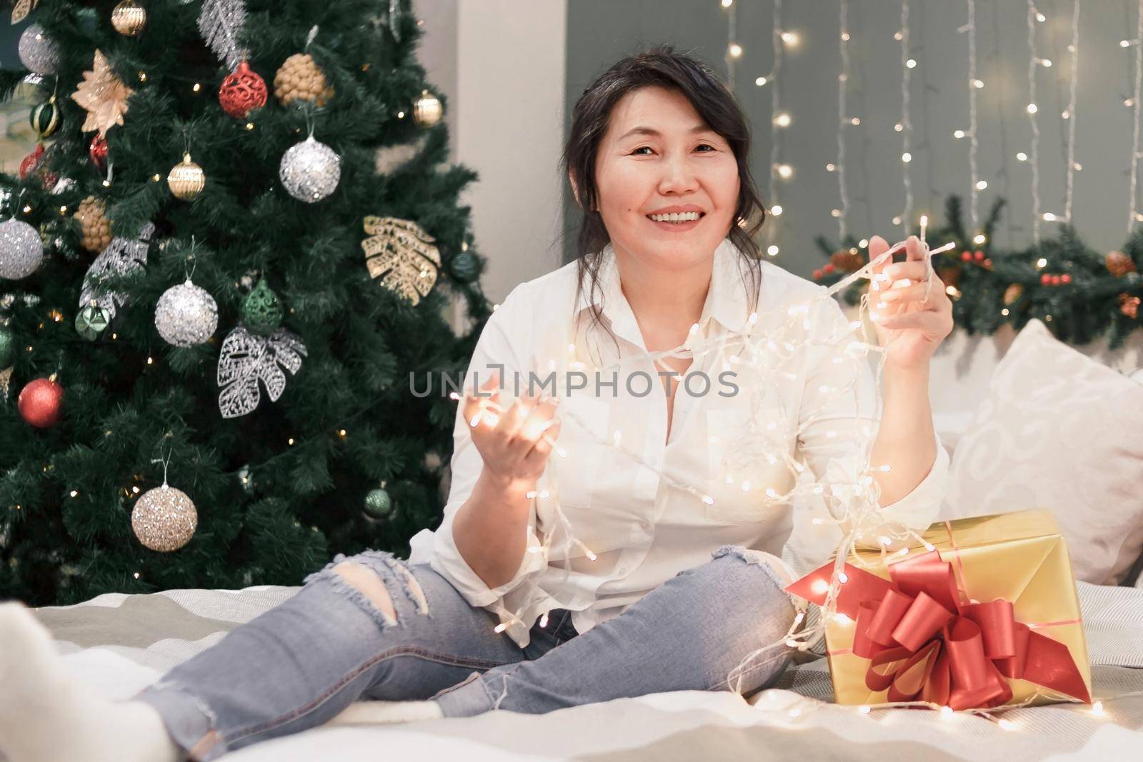 Senior happy asian woman on bed with xmas lights looking at camera, portrait of attractive elderly smiling lady with garland in hands and gold gift at home at christmas holiday, atmospheric mood
