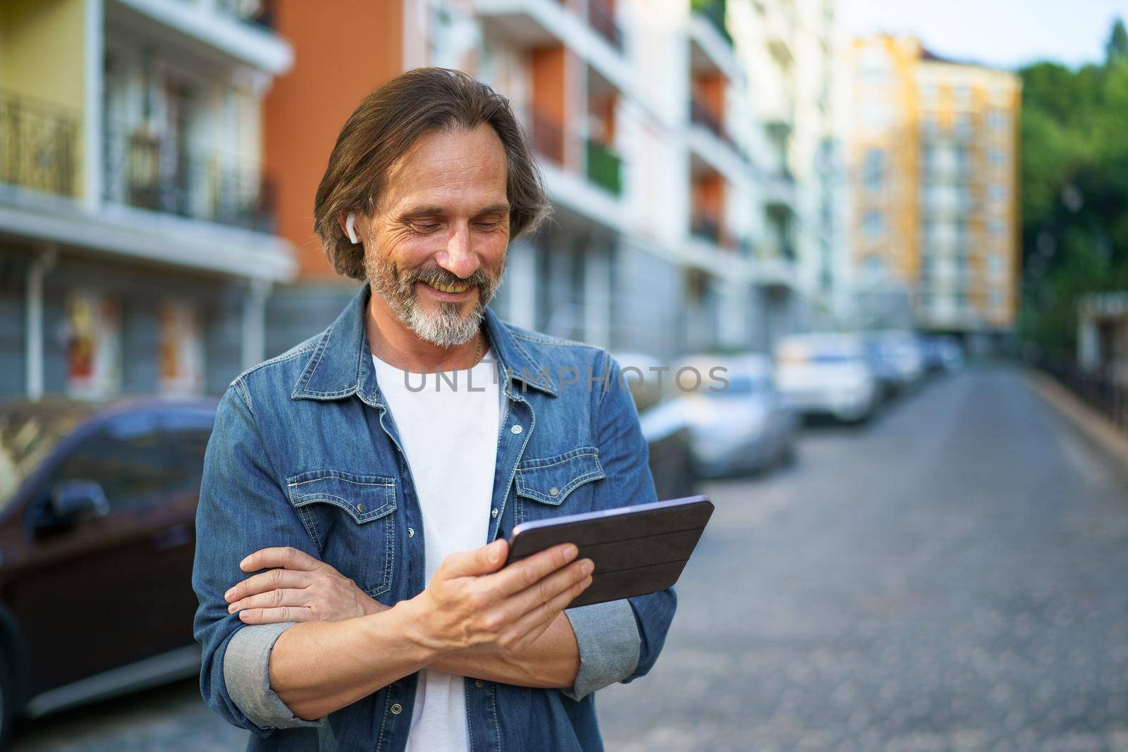 Traveling handsome mature man looking at digital tablet in hand talking outdoors having a call on urban city streets. Mature man listening music use wireless earphones while travel old town streets.