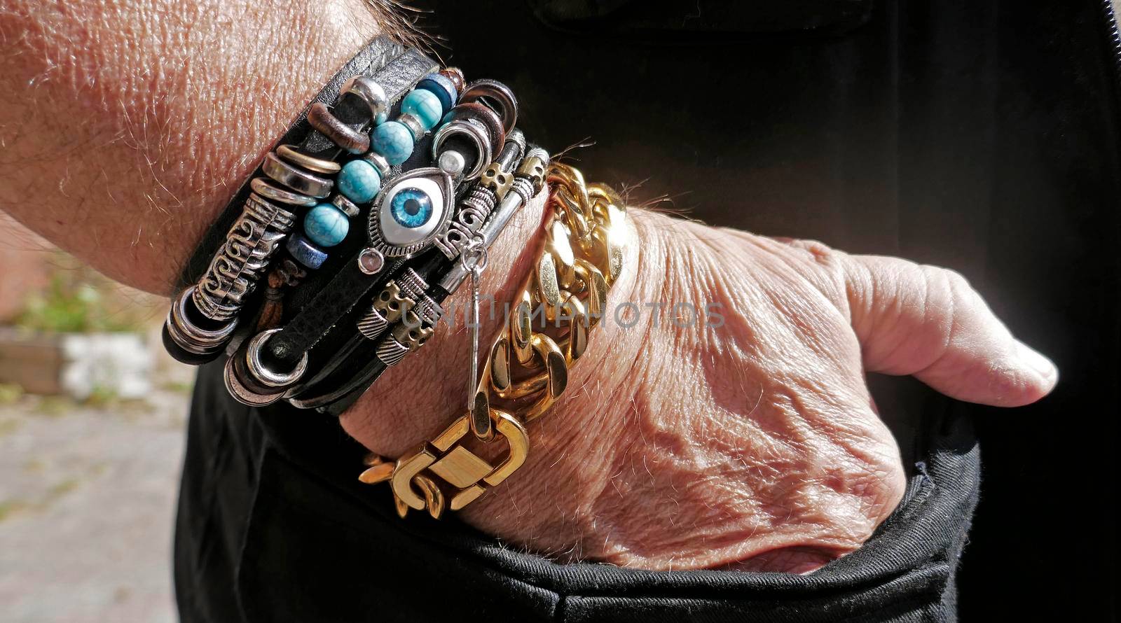 Senior man putting his hand in his pocket. He is wearing a mixture of different bracelets. A large gold colored cuban link bracelet, followed by a group of leather ones