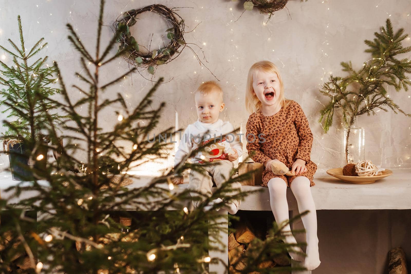 Little brother and sister play on Christmas eve in a beautiful house decorated for the New Year holidays. Children are playing with a Christmas gift. Scandinavian-style interior with live fir trees and a wooden staircase.