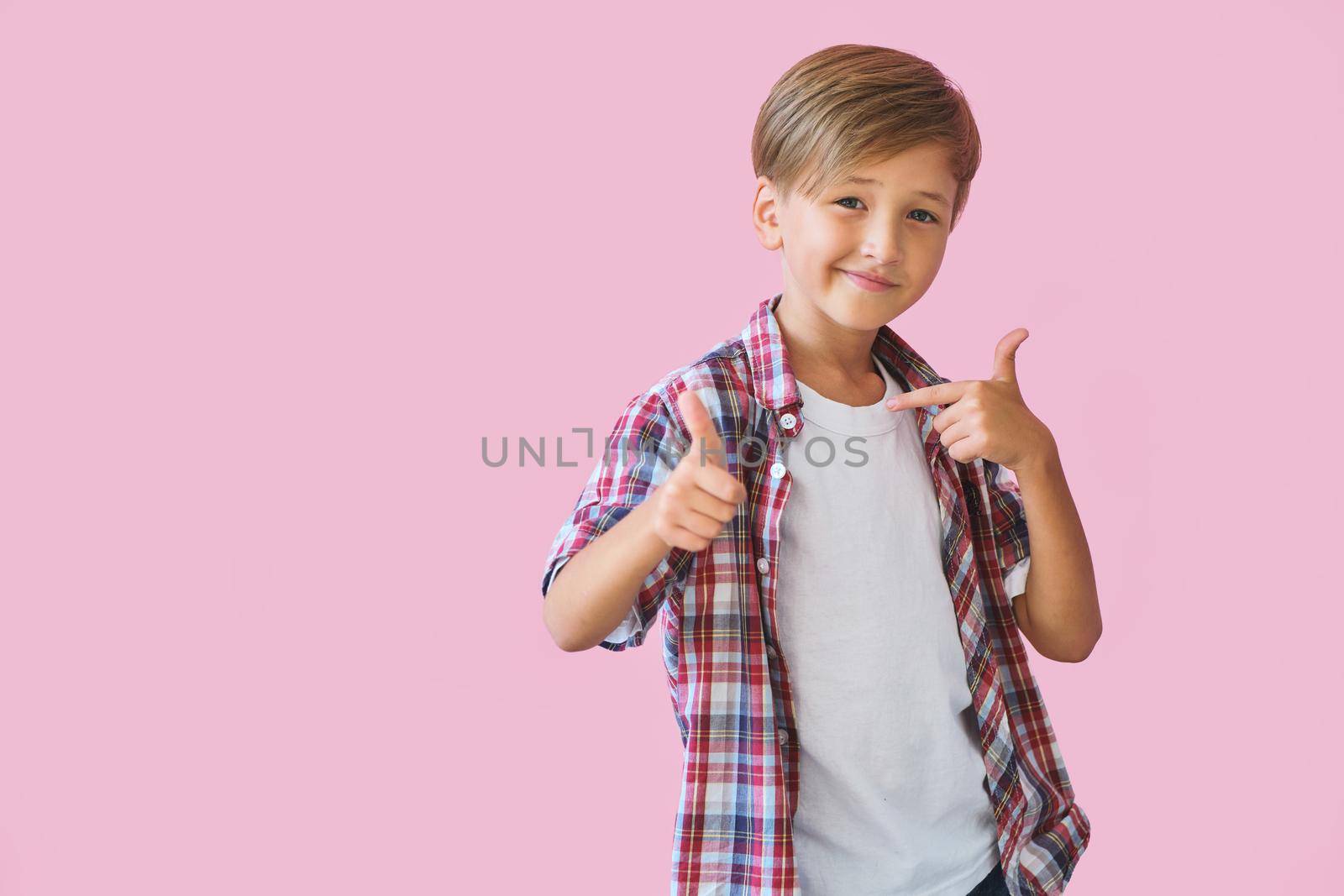 Young happy boy points to his clothes with thumb up, isolated on pink background with copy space. Blank white t-shirt for your design