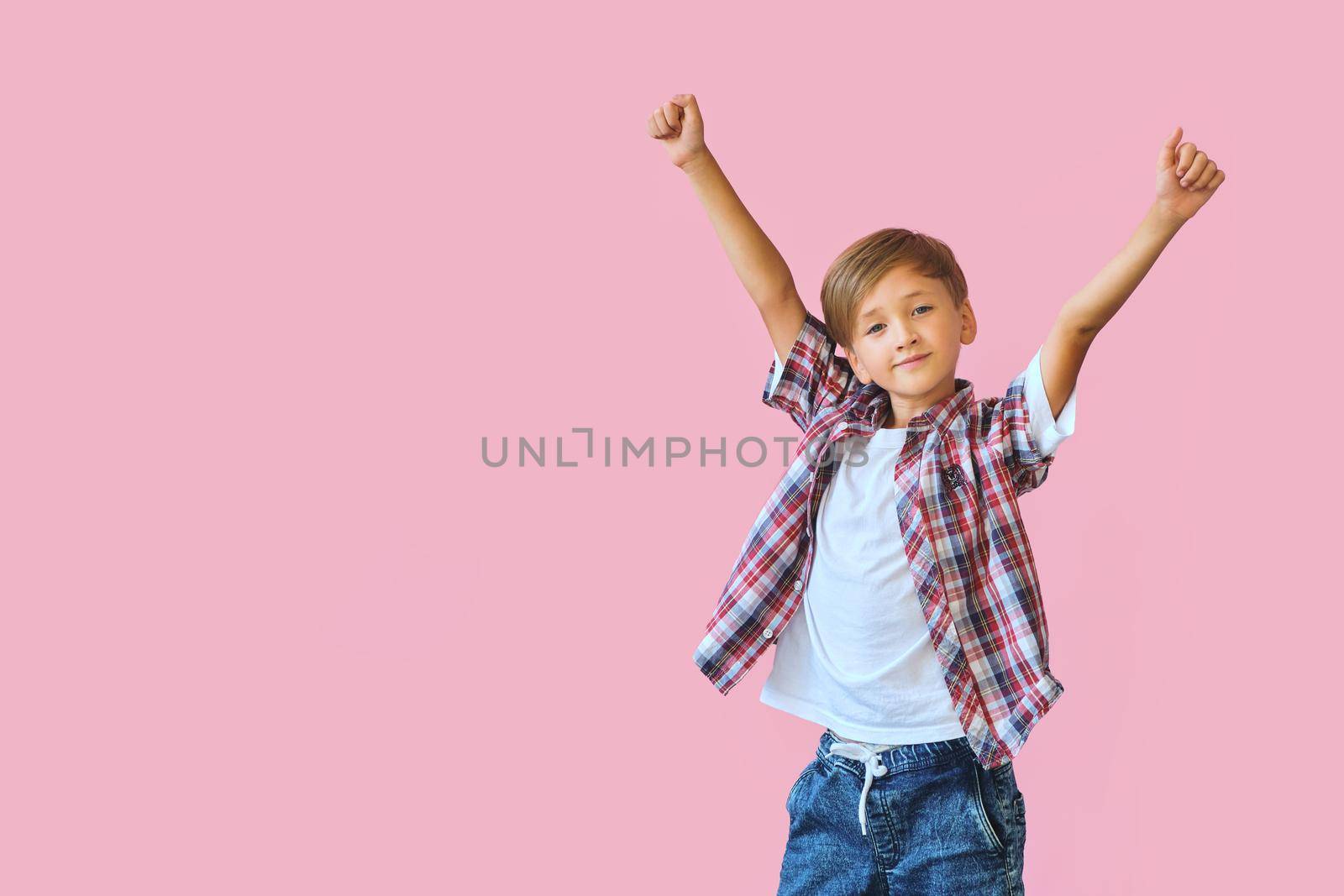 Young happy boy dressed in jeans, a white T-shirt and a plaid shirt with raised hands up isolated on pink background with copy space. Blank white t-shirt for your design