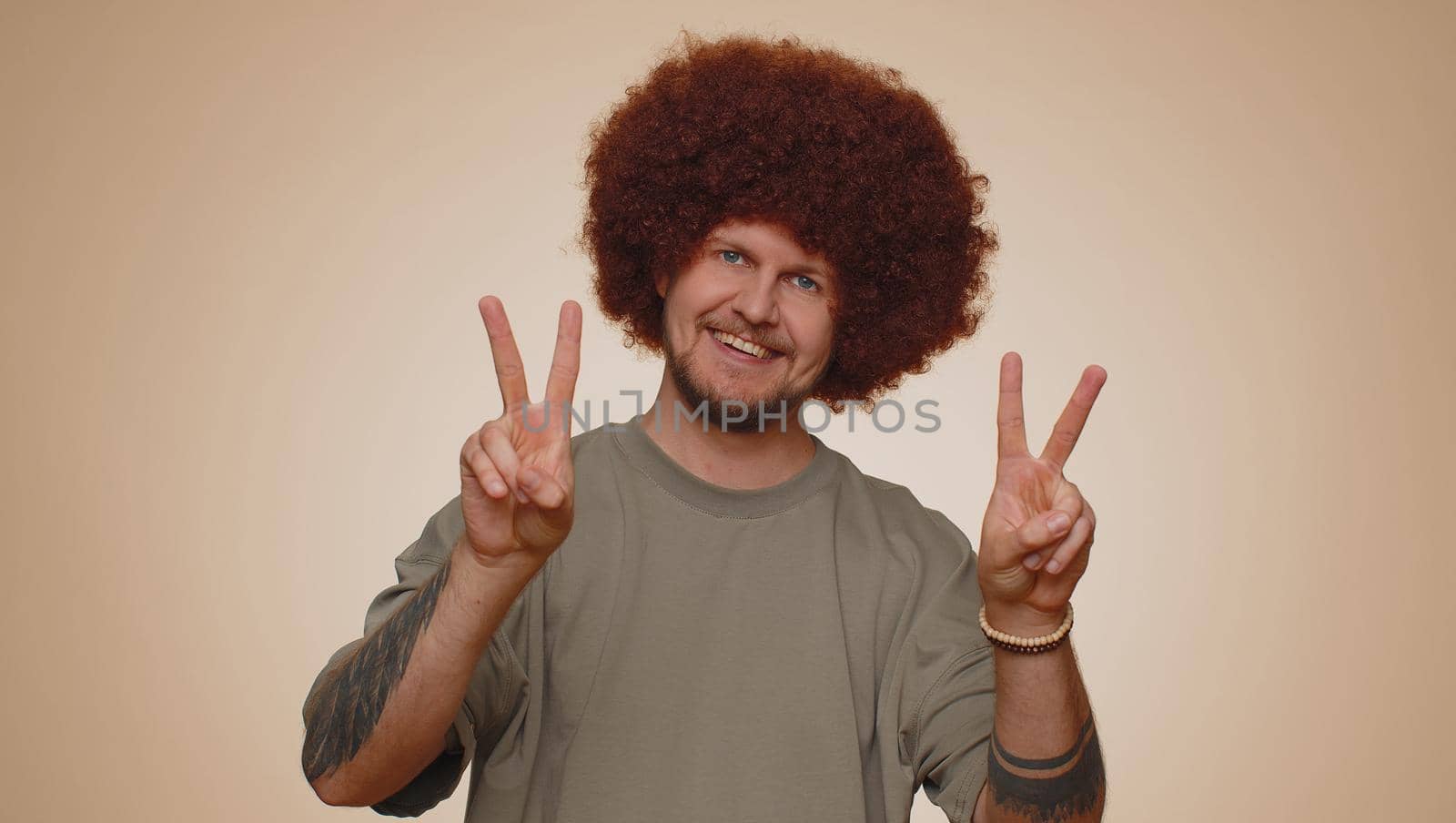 Bearded man with lush hairstyle showing victory sign, hoping for success and win, doing peace gesture, smiling with kind optimistic expression. Young adult guy boy isolated on beige studio background