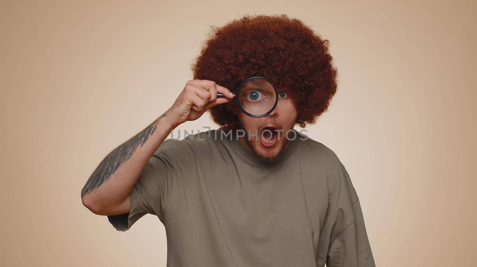 Investigator researcher scientist man holding magnifying glass near face, looking into camera with big zoomed funny eyes, searching, analysing. Young adult guy isolated on beige studio background
