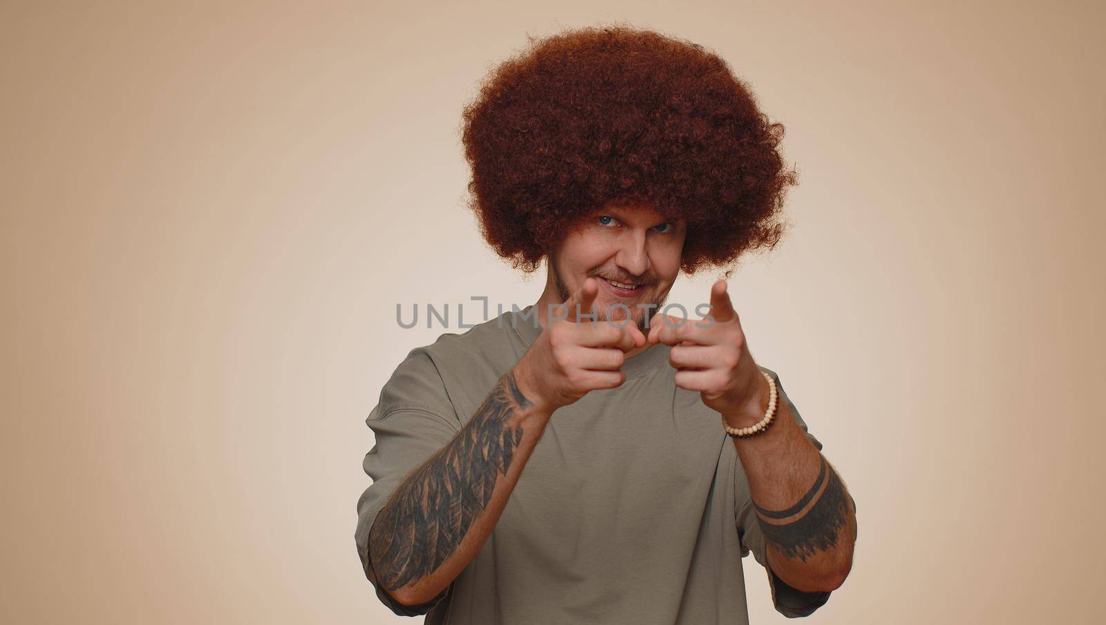 I choose you. Bearded man with lush coiffure pointing to camera and looking with playful happy expression, making choice, showing direction. Young adult guy boy isolated on beige studio background