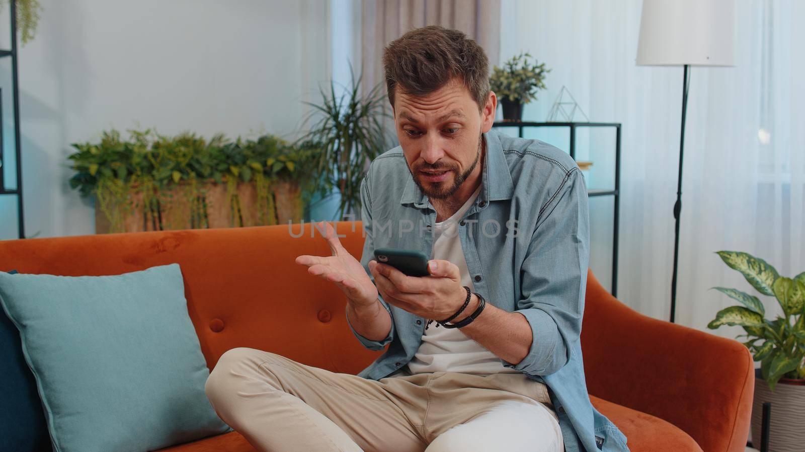 Portrait of caucasian adult man use smartphone typing browsing, loses becoming surprised sudden lottery results, bad news, fortune loss, fail. Young guy sitting on orange couch at home living room