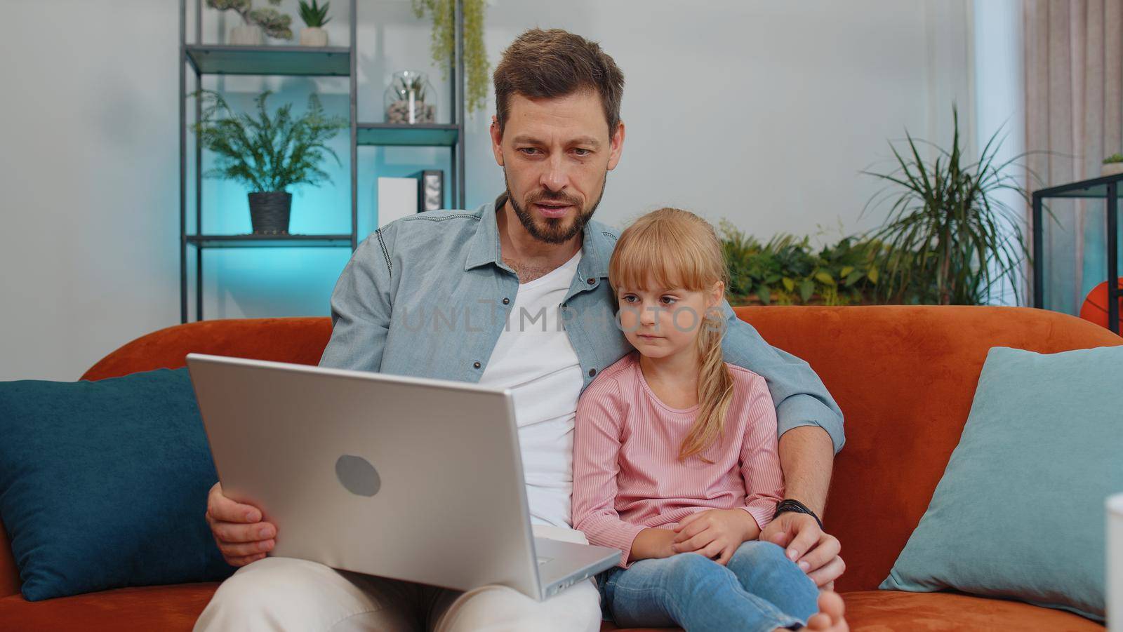 Happy family father or nanny and child kid daughter laugh use laptop, watch cartoons, make video call, look at screen. Dad with child making online shopping on notebook sit on sofa at home living room