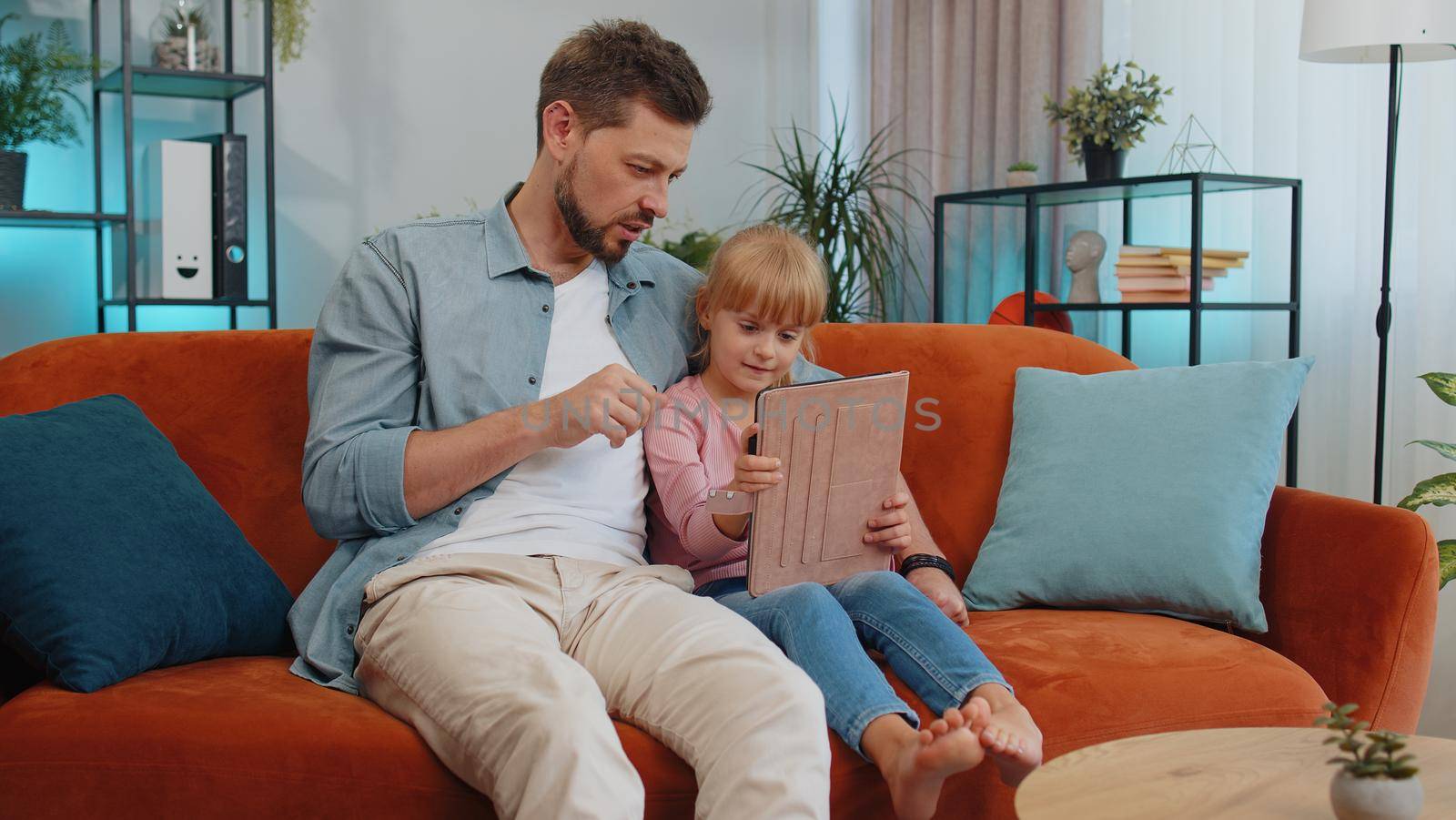 Happy family father or nanny and child kid daughter laugh use digital tablet, watch cartoons, make video call, play game, study. Dad with child making online shopping sit on sofa at home living room