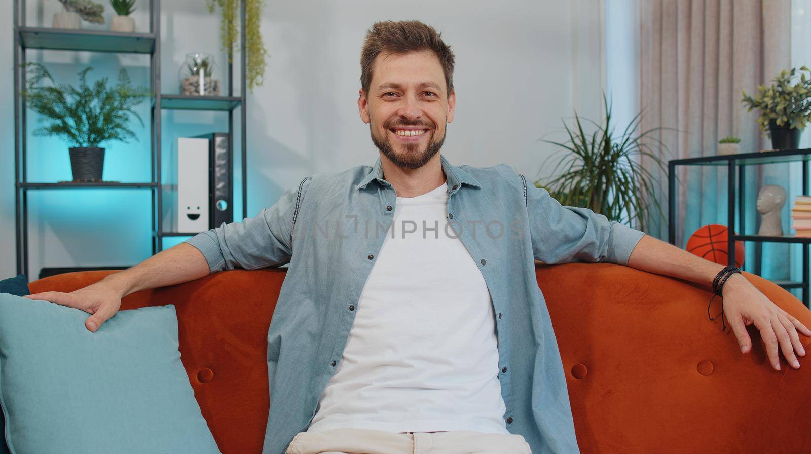 Close-up portrait of happy smiling caucasian adult man in shirt, looking at camera, celebrate good news. Young guy indoors isolated at home in living room sitting on orange couch. Male nature beauty