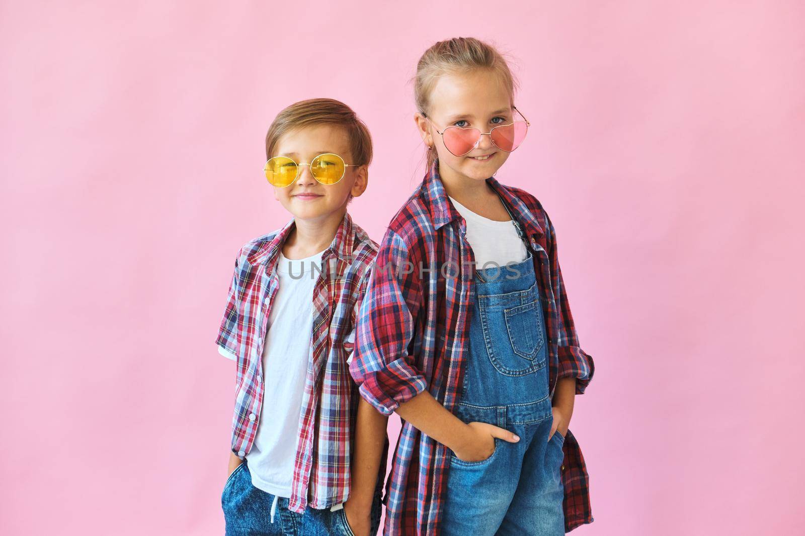 Cute stylish little girl and boy in color sunglasses dressed in plaid shirts, looking at camera and laughing while standing on pink background with copy space.