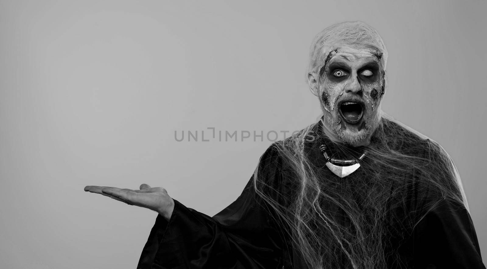 Zombie man with makeup with fake wounds scars showing thumbs up and pointing empty place, advertising area for commercial text logo, copy-space for goods promotion. Sinister Halloween ugly dead guy